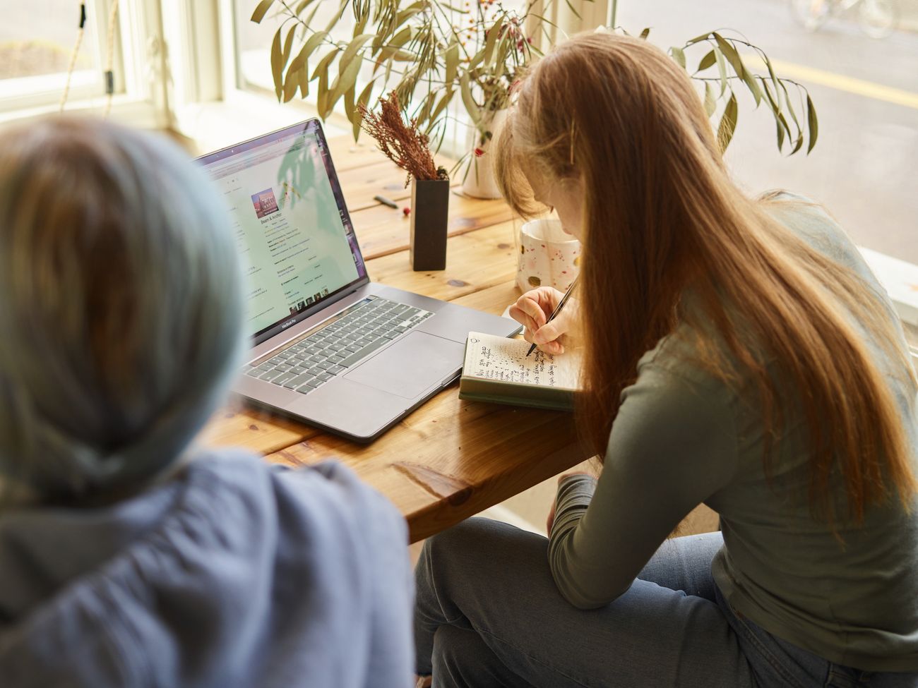 A person taking notes in a notebook while researching on a laptop.